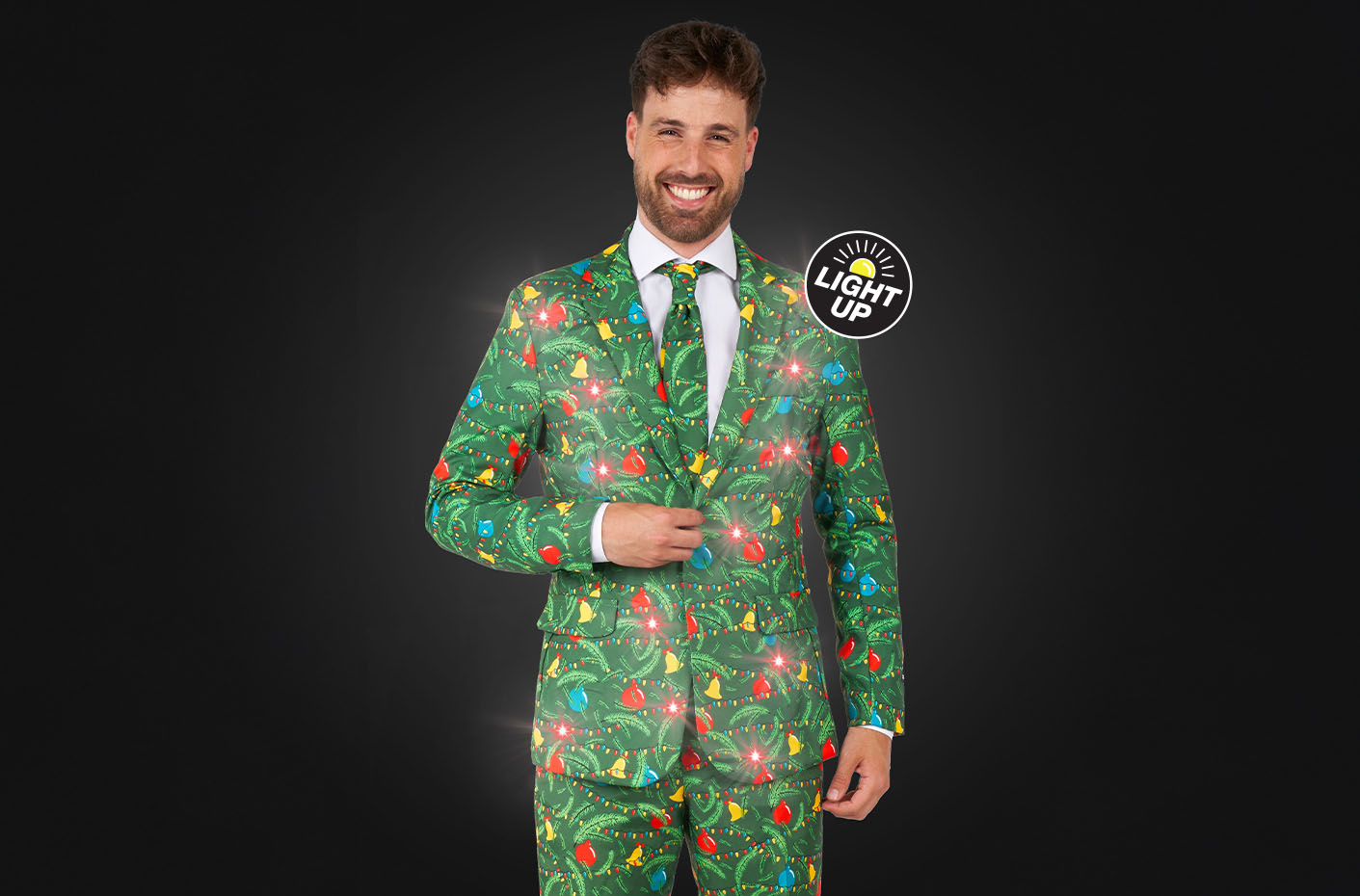 Image of a man wearing a green Christmas suit with lights, standing in front of a dark background, smilling at the camera. Suits consist of a blazer, pants and tie.
