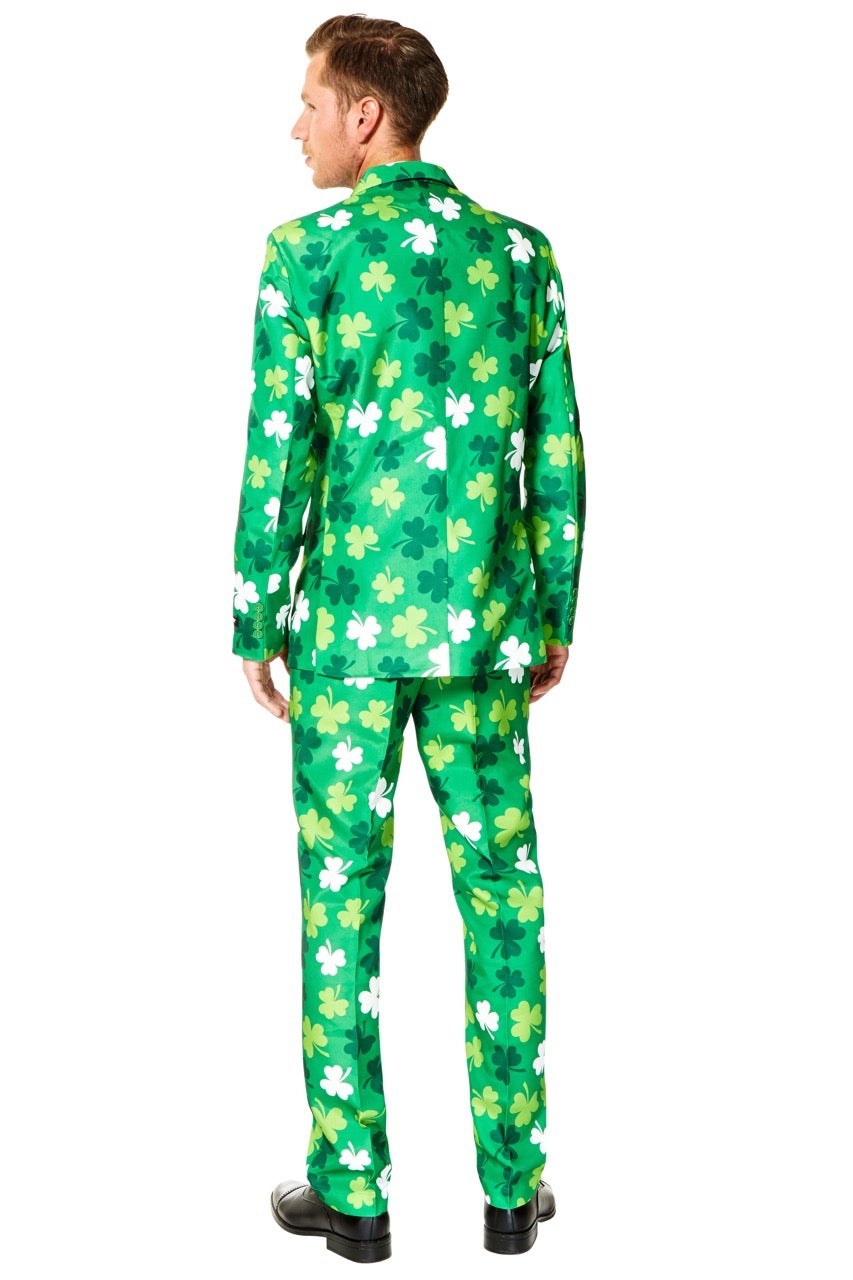 A man in a bright Suitmeister ensemble, featuring the St Patricks Day Clovers design with white and dark green shamrock patterns, stands facing away. He has short hair and wears black shoes, completing his festive look with a matching jacket and pants.