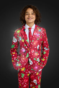 A smiling child with curly hair wears the Suitmeister BOYS Christmas Red Icons - Light Up suit, featuring snowmen, Christmas trees, and gifts that sparkle with glowing lights against a dark background.
