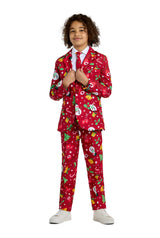 A child smiles while holding the lapels of the Suitmeister BOYS Christmas Red Icons - Light Up suit, featuring festive snowmen, trees, and ornaments. He pairs it with a white shirt, red tie, and white sneakers.