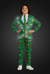 A smiling child stands hands on hips, wearing the Suitmeister BOYS Christmas Green Tree - Light Up suit, decorated with festive lights, trees, and ornaments. The suit appears to sparkle against a dark gray background.