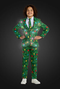 A smiling child stands hands on hips, wearing the Suitmeister BOYS Christmas Green Tree - Light Up suit, decorated with festive lights, trees, and ornaments. The suit appears to sparkle against a dark gray background.