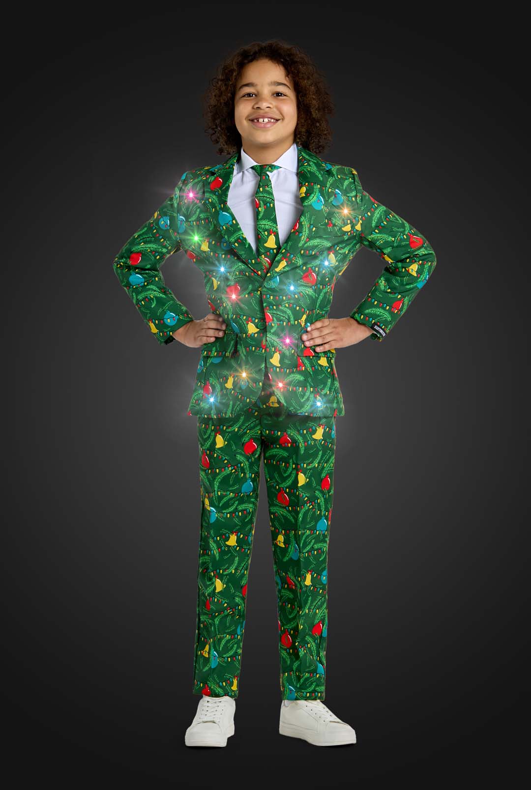 A smiling child stands hands on hips, wearing the Suitmeister BOYS Christmas Green Tree - Light Up suit, decorated with festive lights, trees, and ornaments. The suit appears to sparkle against a dark gray background.