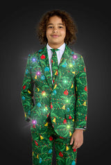 A boy with curly hair wears the Suitmeister BOYS Christmas Green Tree - Light Up suit, adorned with colorful ornaments and lights, standing against a dark background. The festive green outfit sparkles brightly.