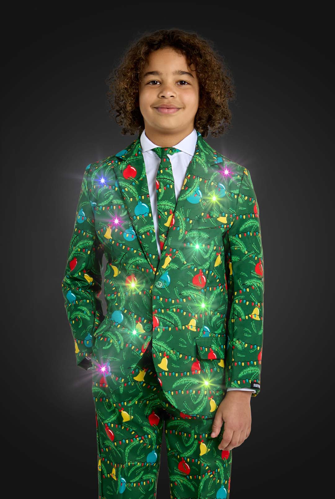 A boy with curly hair wears the Suitmeister BOYS Christmas Green Tree - Light Up suit, adorned with colorful ornaments and lights, standing against a dark background. The festive green outfit sparkles brightly.