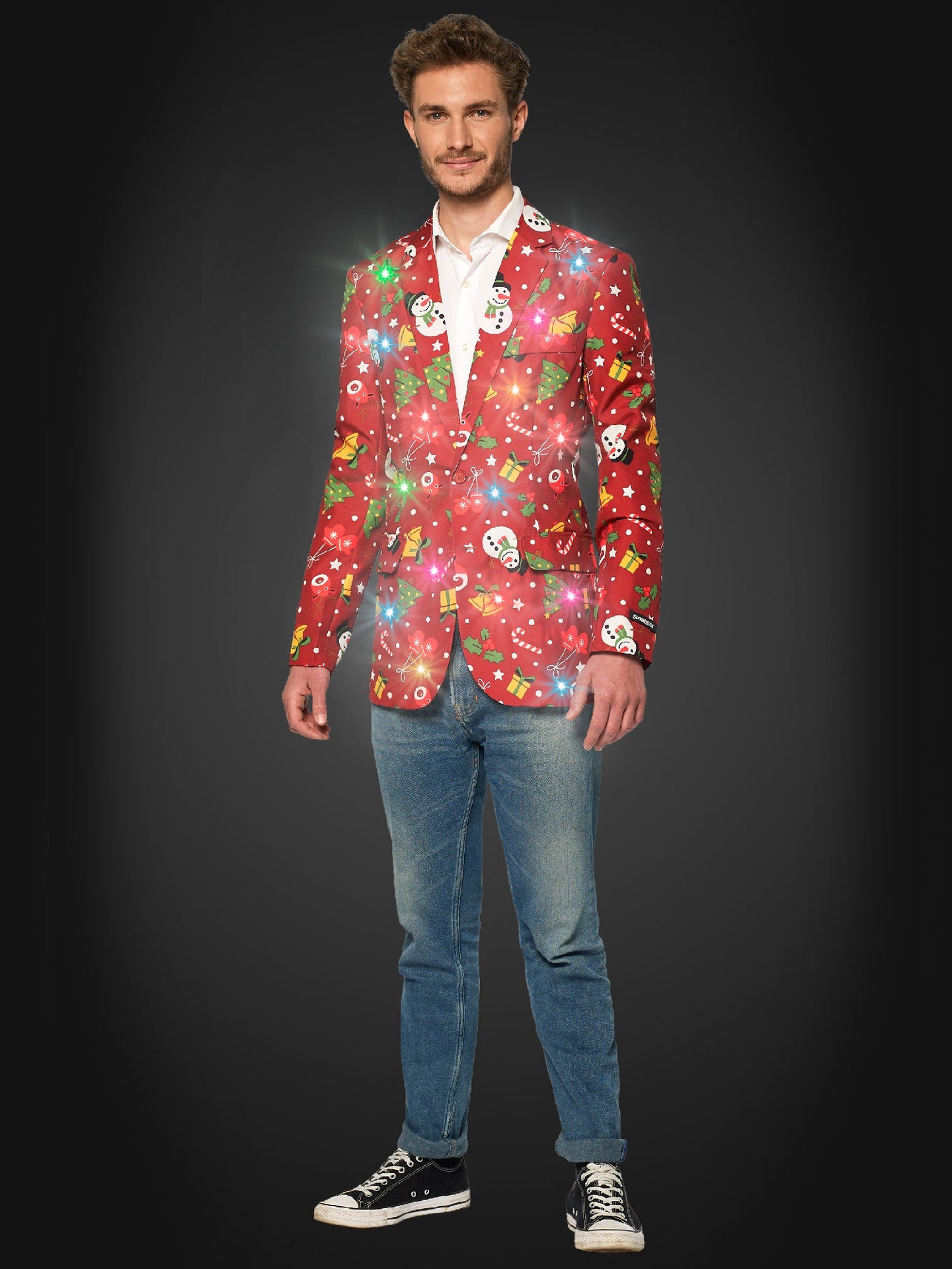 A man smiles, wearing the Suitmeister "Christmas Red Icons - Light Up" blazer with festive patterns and glowing lights, paired with a white shirt, blue jeans, and black sneakers against a dark background.
