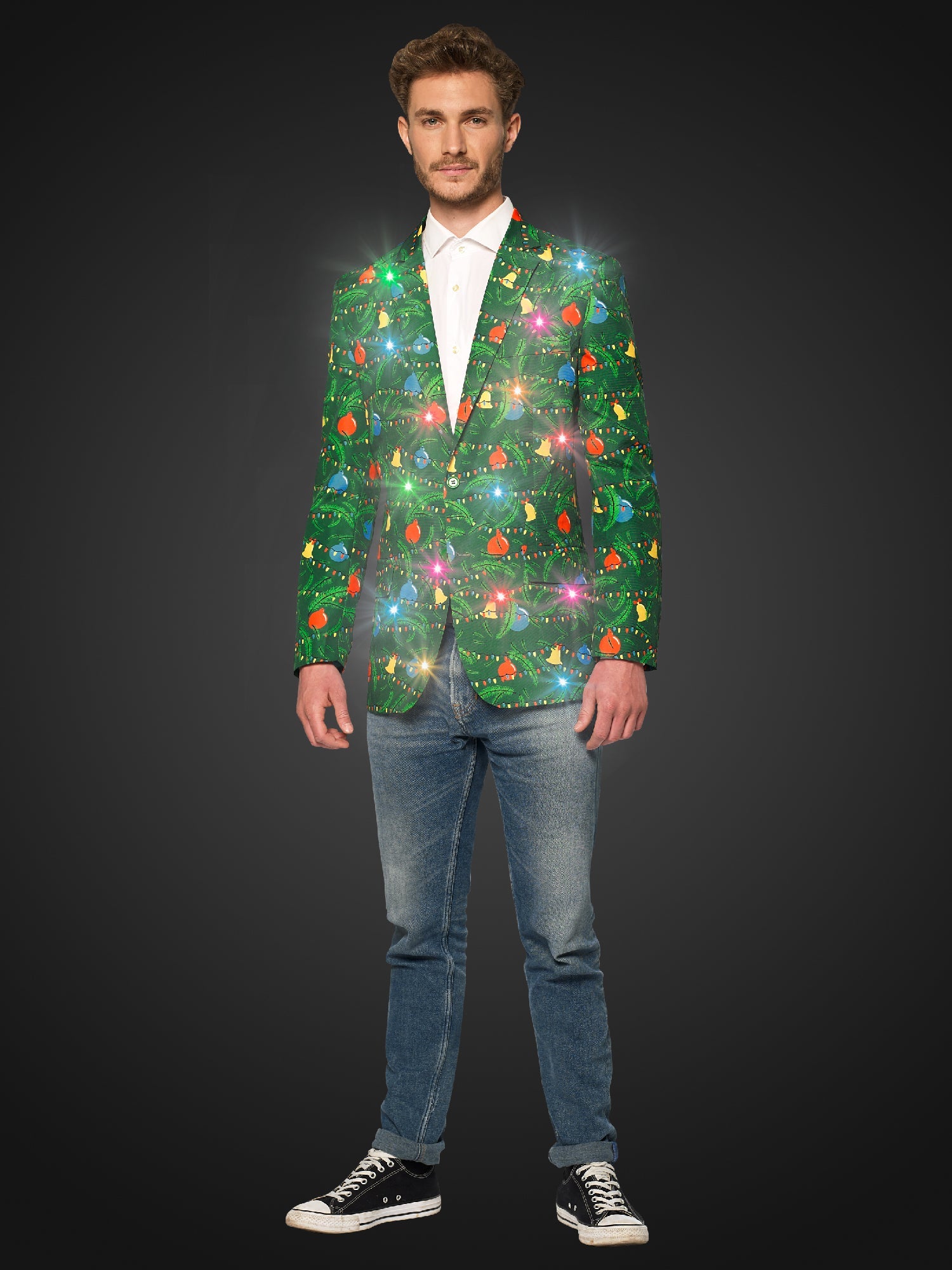 A man stands against a dark background wearing blue jeans, black sneakers, and the Suitmeister Christmas Green Tree - Light Up blazer, decorated with glowing Christmas lights and ornaments.