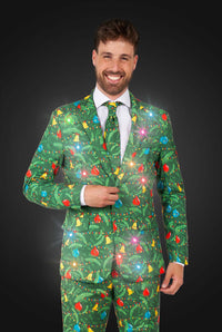 A man smiles in the dark, wearing the Suitmeister Christmas Green Tree - Light Up suit, adorned with colorful ornaments and glowing lights.