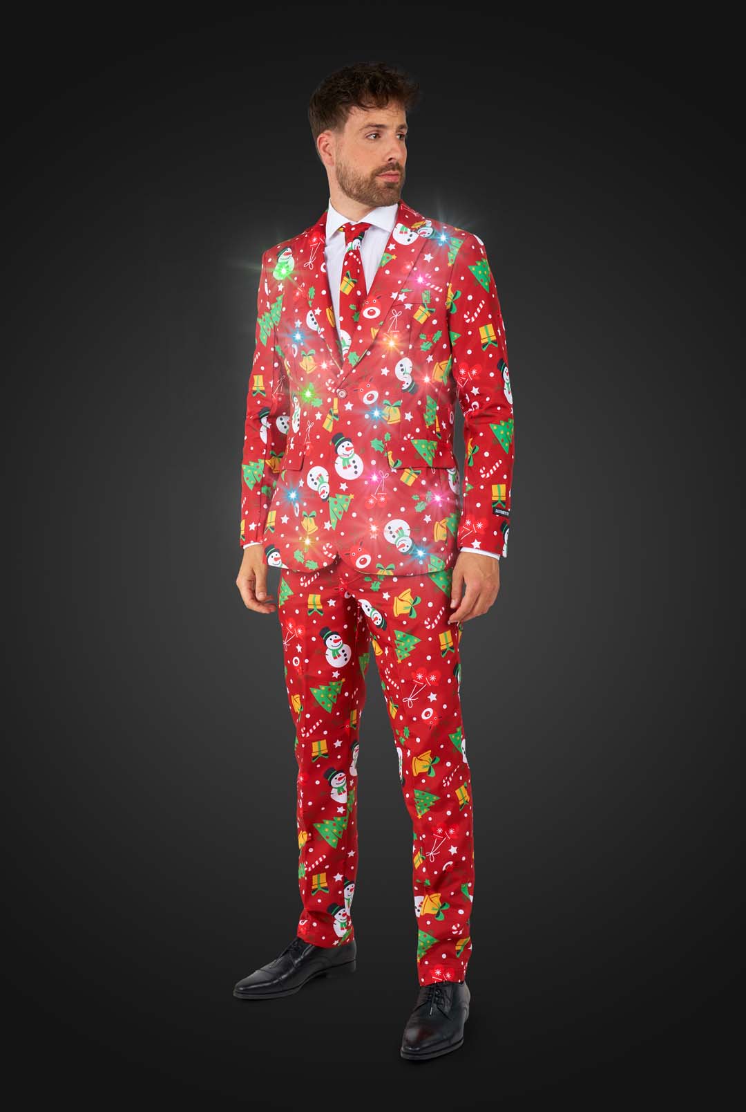 A man in a Suitmeister Christmas Red Icons - Light Up suit featuring snowmen, gifts, and stars stands against a dark background. The glowing red suit is paired with a white shirt, black shoes, and a tie for a festive formal look.