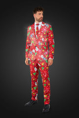 A man in a Suitmeister Christmas Red Icons - Light Up suit featuring snowmen, gifts, and stars stands against a dark background. The glowing red suit is paired with a white shirt, black shoes, and a tie for a festive formal look.