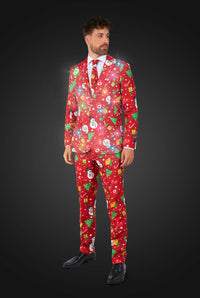A man in a Suitmeister Christmas Red Icons - Light Up suit featuring snowmen, gifts, and stars stands against a dark background. The glowing red suit is paired with a white shirt, black shoes, and a tie for a festive formal look.