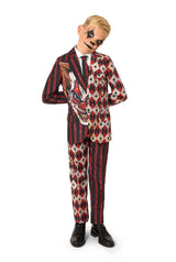 Full-body view of a child in a red and black patterned clown suit with a white shirt and black tie, standing with hands behind their back.