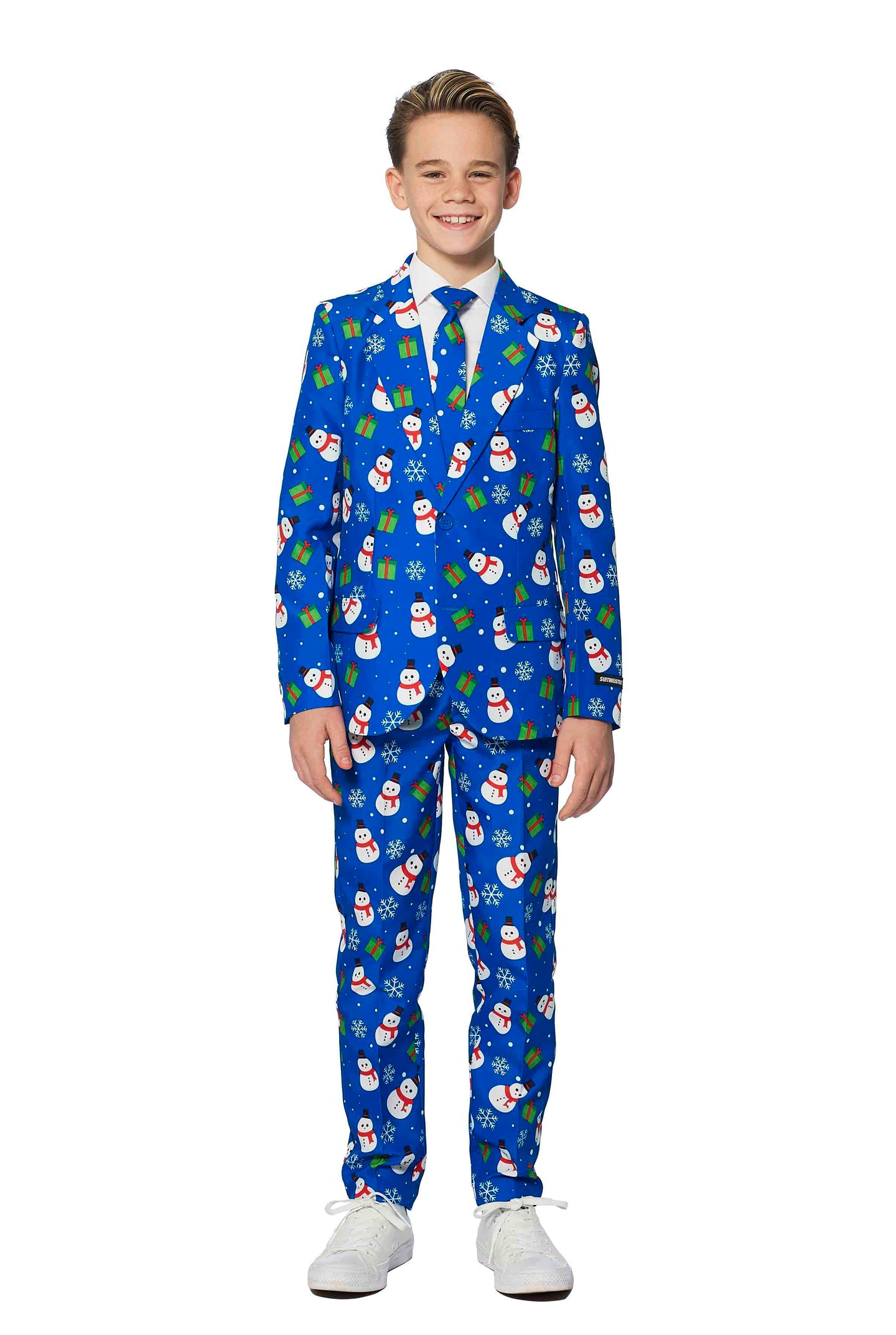 A young boy is beaming with joy in a Suitmeister's Christmas Suit Blue Snowman. The festive attire features a cheerful Christmas-themed pattern adorned with snowmen, holly, and presents. He stands against a plain white background while wearing white shoes.