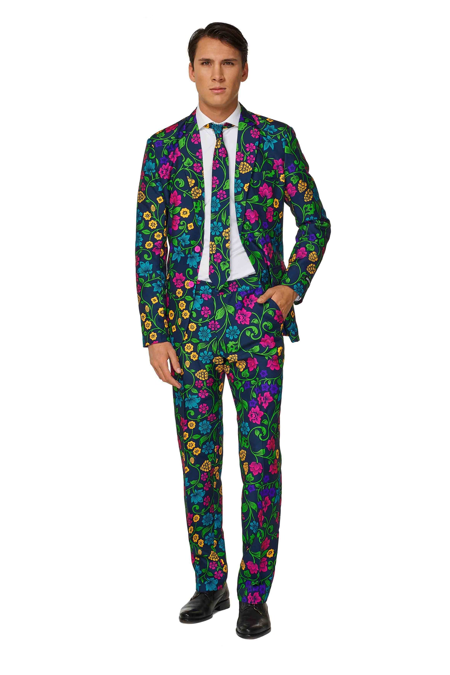 A man wearing the Suitmeister Floral, a vibrant suit adorned with a colorful floral pattern featuring blue, pink, green, and yellow flowers. He complements it with a white shirt, black tie, and black shoes while standing against a plain white backdrop.