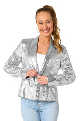 A smiling woman with long, light brown hair in a ponytail wears the Suitmeister Sequins Silver sequin blazer over a white top and blue jeans, holding the blazer closed with both hands against a white background.